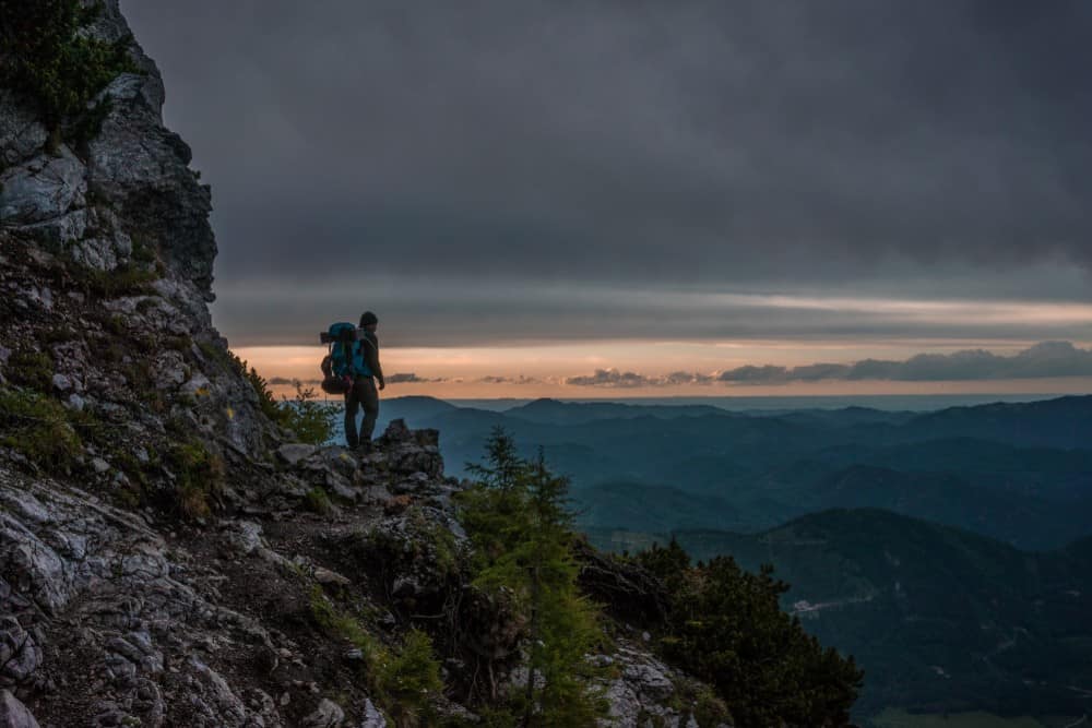 Ein Mann steht bei Sonnenuntergang auf einem Berggipfel in Österreich-Urlaub und bewundert mit seinen Kindern die atemberaubende Aussicht.